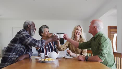 Two-diverse-senior-couples-sitting-by-a-table-drinking-tea-together-making-a-toast-at-home