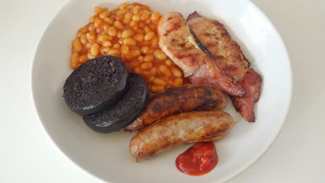 tomato ketchup being added to a traditional english breakfast of cumberland sausages, back bacon black pudding and baked beans, but no egg, on a white plate