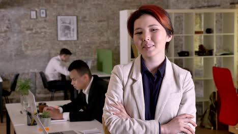 young business asian woman with crossed hands looking in camera in modern office, smiling, black and caucasian mans working in back