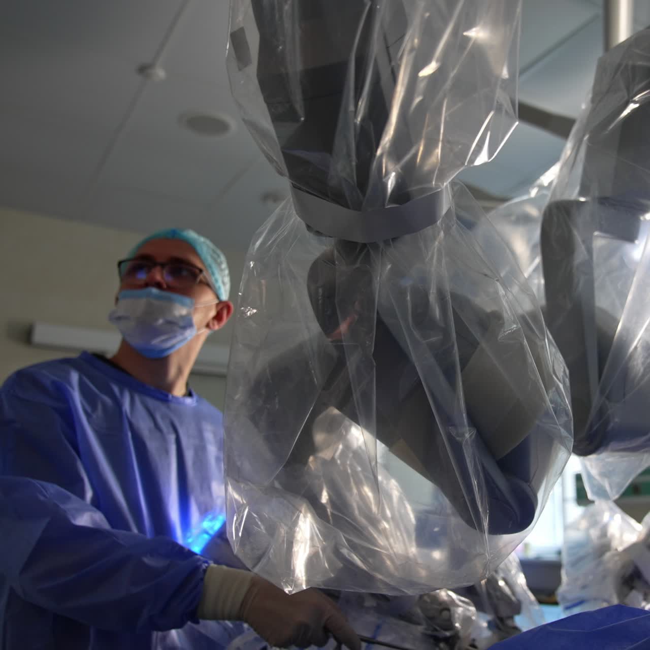 Sterile covered robotic arms performing the operation in modern surgery room. Doctor in glasses standing beside holding the tool