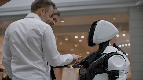 two people a man and a woman communicate with a robot. press the robot with your hands on the screen. a robot assistant interacting in a shopping center with a couple. robot assistant in the trading floor