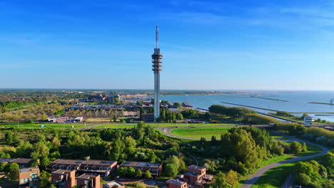 flying closer to the telecom tower in lelystad, the netherlands. aerial perspective on the panorama of green city on sunny day. lake merkemeer with dams at backdrop.