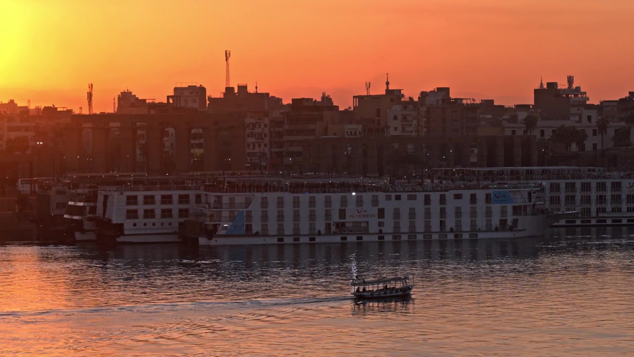 Lo skyline di Luxor illuminato da un brillante tramonto arancione, con navi da crociera ormeggiate lungo la Sponda Orientale e un motoscafo tradizionale che naviga le acque scintillanti del Nilo.