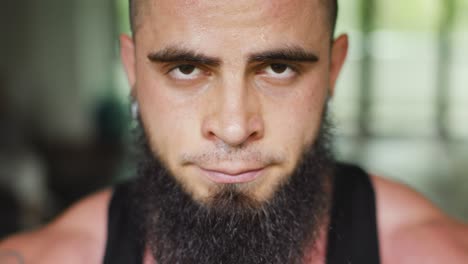 Young-man-with-beard-looks-into-the-camera,-sweating-and-breathing-strong-after-exercise-in-gym-close-up