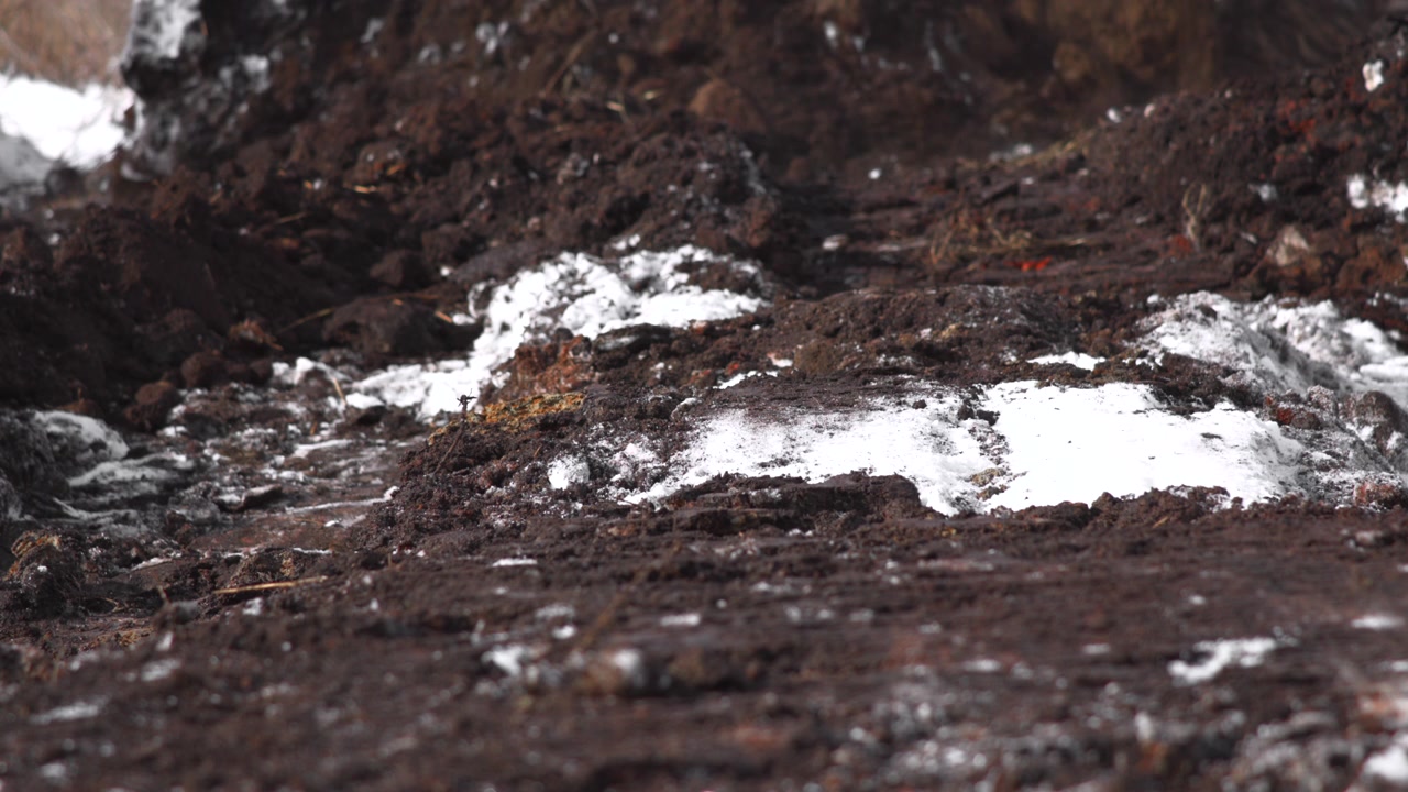 Primo piano ruvido di zolle di terra scura o rocce con chiazze sparse di neve e brina, visto da un punto di vista molto basso con profondità di campo ridotta, toni smorzati di nero, grigio e bianco, superficie strutturata e freddo.