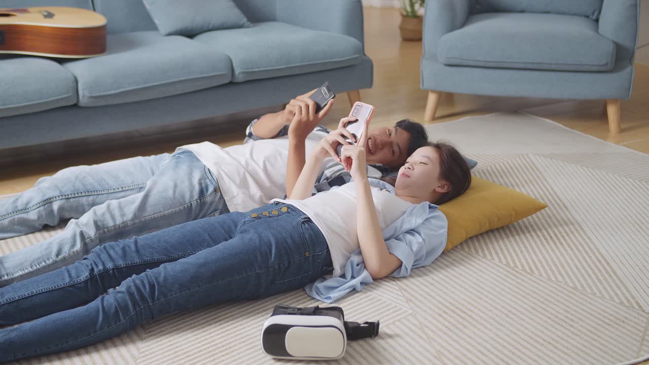 아시안 십대 커플이 집 바닥에 카에 누워 스마트폰을 연주하고 웃고, 이야기하고, 함께 시간을 즐기고 있습니다.