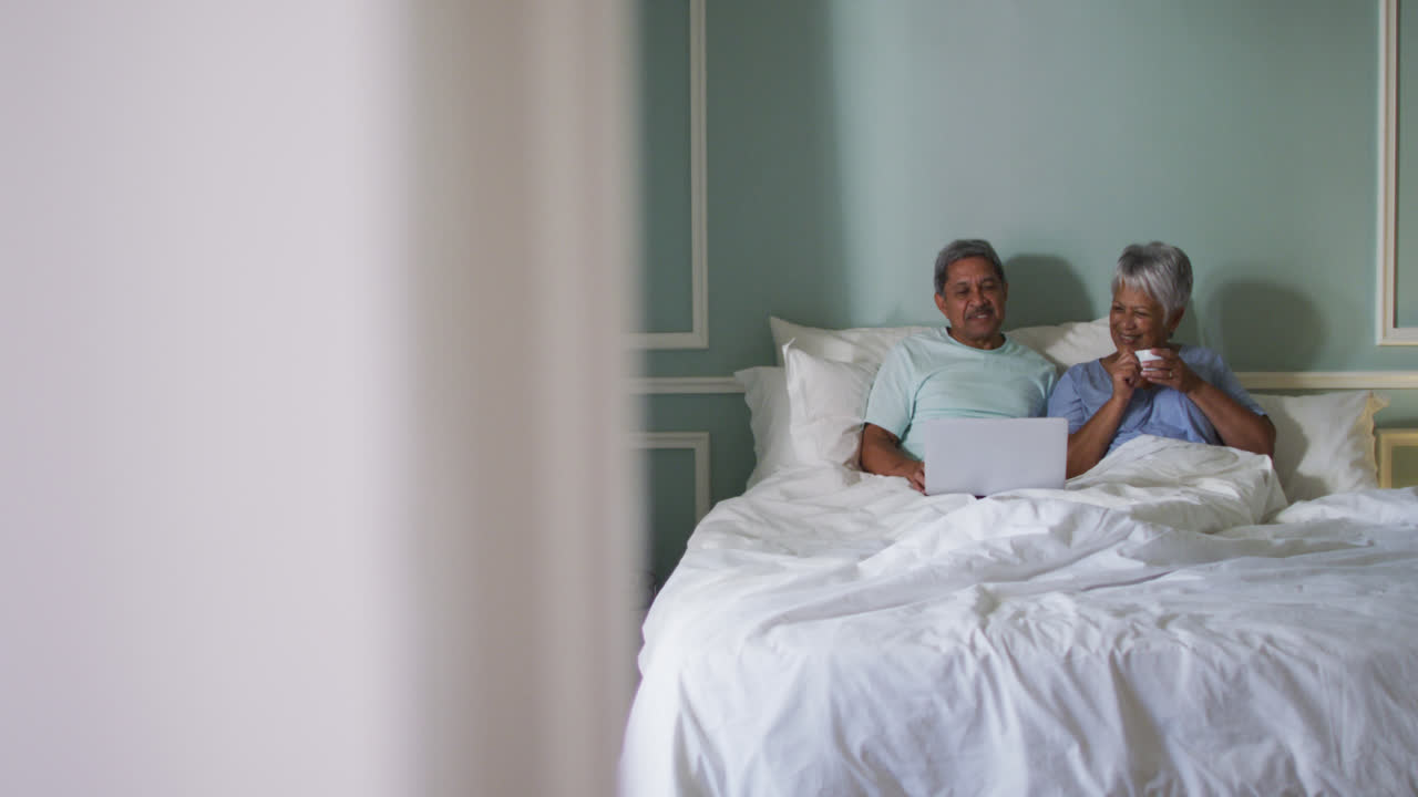 Happy senior mixed race couple sitting in bed using laptop