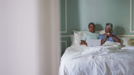 Happy-senior-mixed-race-couple-sitting-in-bed-using-laptop