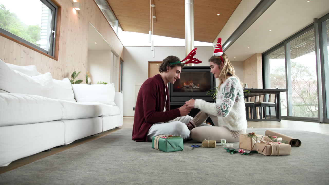 Couple wrapping Christmas gifts at home, smiling by cozy fireplace