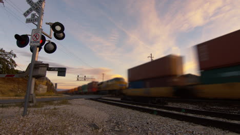 a super sped up shot of a really long train passing at a train stop at sunset while the lights of the gate flash.