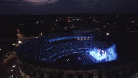 Rotación-De-Drones-A-La-Izquierda-En-Las-Arenas-De-Nîmes-En-Medio-De-La-Noche,-La-Gente-Está-Viendo-El-Concierto-Y-Hay-Luces-De-Varios-Colores