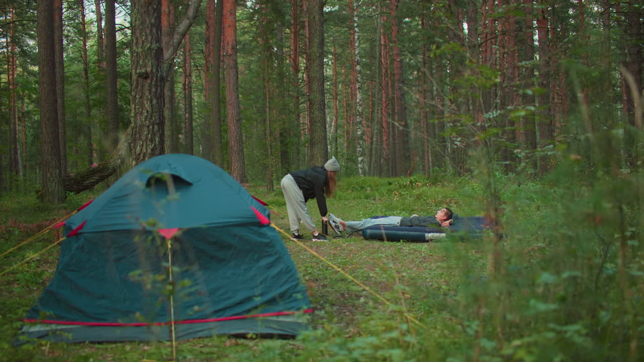 white man lies on air bed while woman pumps air into it with pitched tent behind in forest clearing surrounded by trees and soft daylight