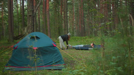 white man lies on air bed while woman pumps air into it with pitched tent behind in forest clearing surrounded by trees and soft daylight