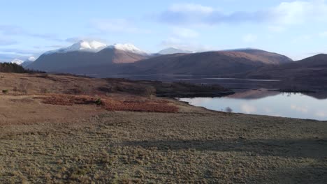 Aerial-drone-footage-flying-close-above-moorland-and-fields-towards-a-stunning-view-of-Glen-Etive-and-Loch-Etive-in-the-Highlands-of-Scotland-with-snow-capped-mountains-and-still-reflective-water