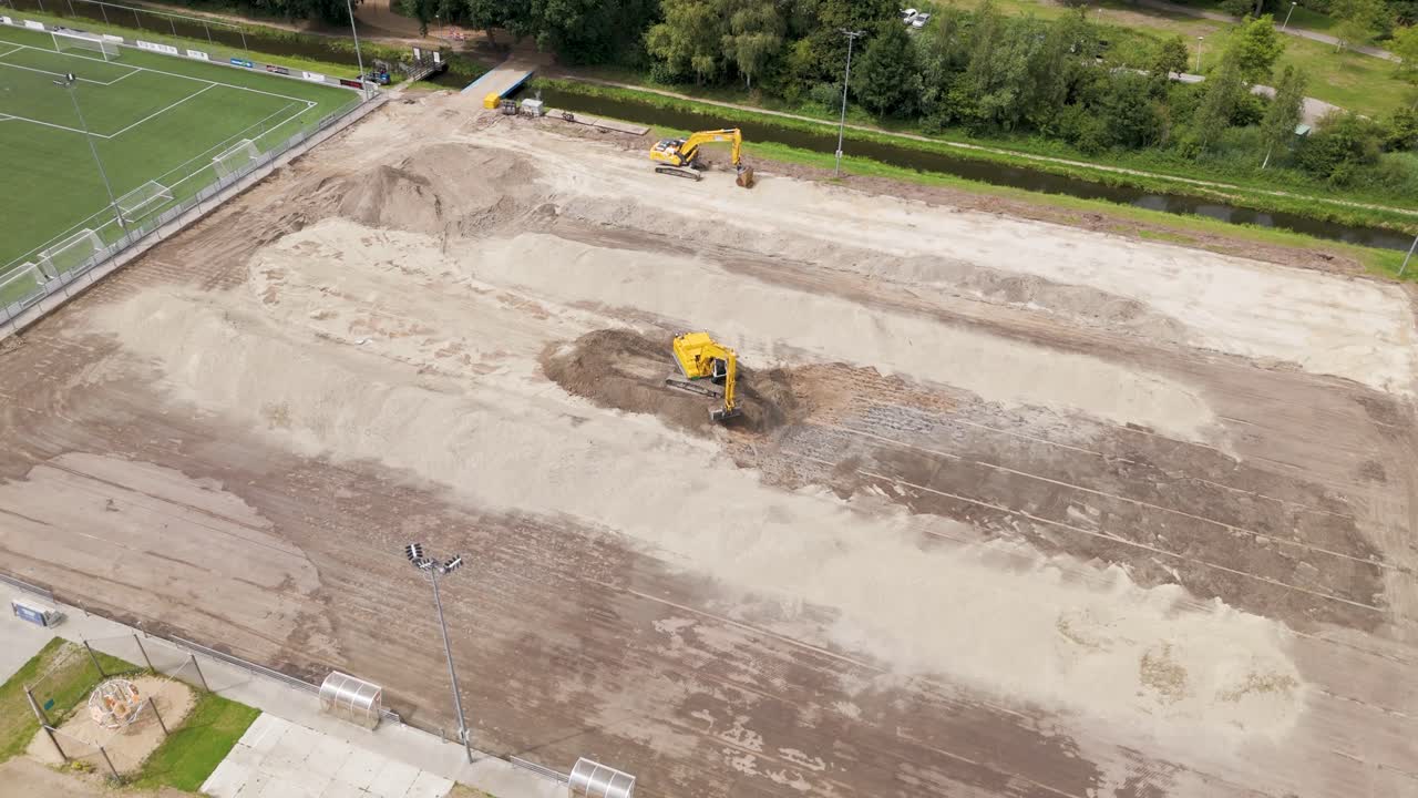Drone footage showing multiple excavators working on the preparation of a large sports field, surrounded by greenery and a canal