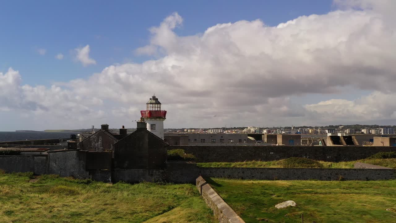 Aerial orbit of Mutton Island’s coastline with historic lighthouse and rock walls across green landscape, rocky shores, and expansive ocean in Galway Bay, Ireland as flock of seagulls soar in sky