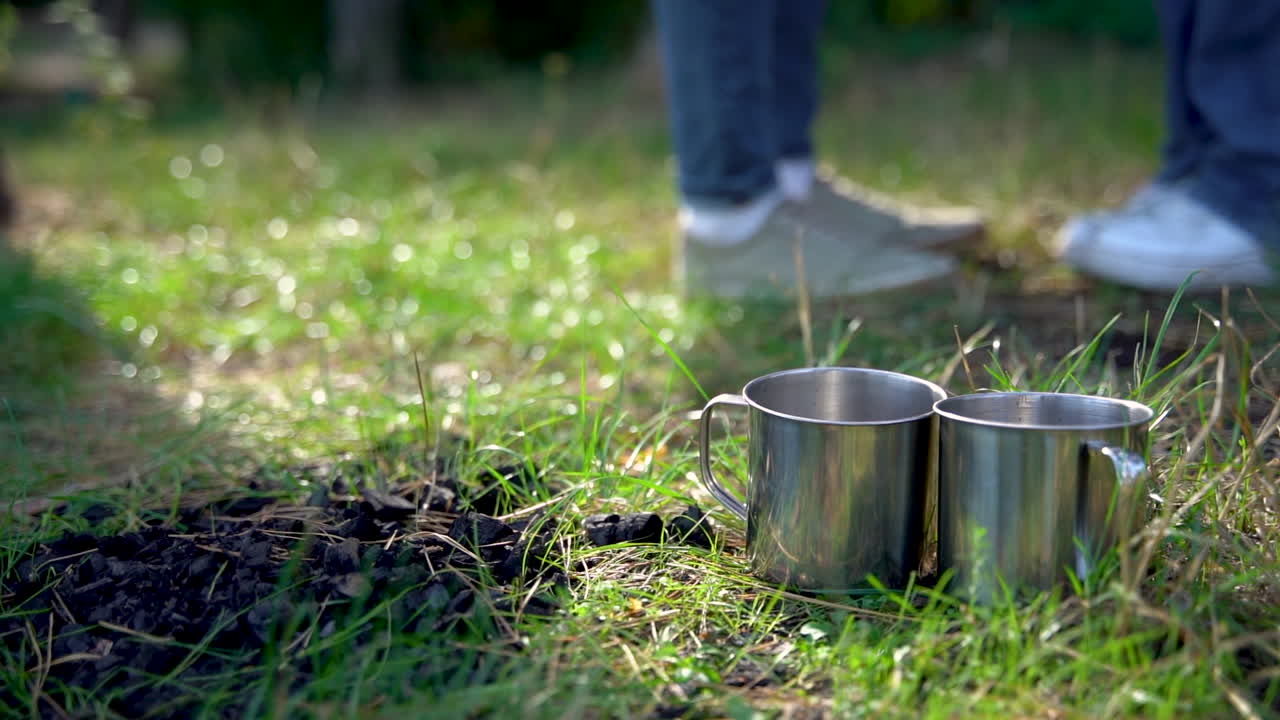Two stainless steel cups on the sunny grass of the woods. Close-up. Camping day in the mountains.