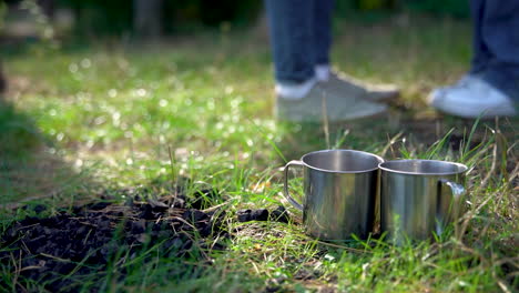 Two-stainless-steel-cups-on-the-sunny-grass-of-the-woods.-Close-up.-Camping-day-in-the-mountains.