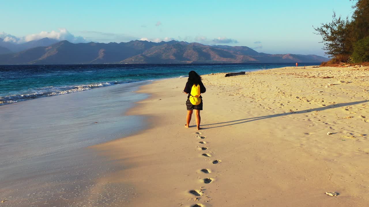 chica con mochila amarilla caminando descalza por una playa exótica bañada por olas marinas al amanecer con colores vivos del paisaje marino en indonesia