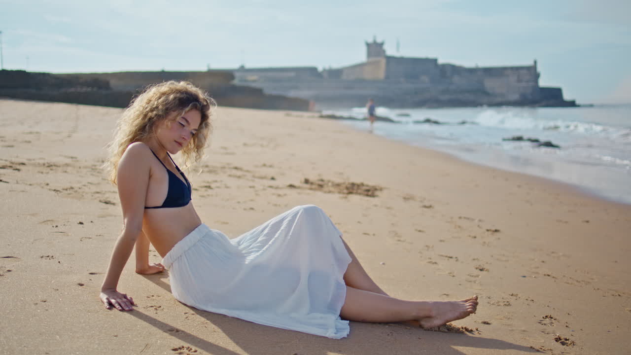 아름다운 관광객이 해변에 앉아있는 은 날 카메라를 보고 있습니다. 모래에서 휴식을 취하는 여성