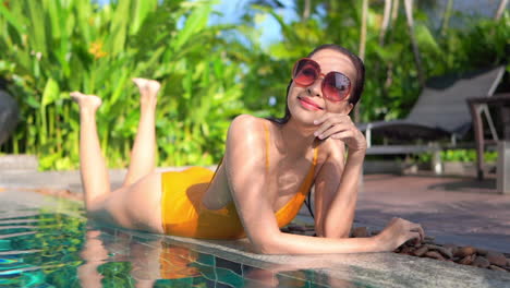 Smiling-Asian-girl-with-big-sunglasses-sunbathes-lying-on-edge-of-pool