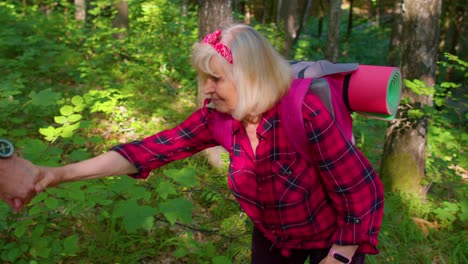 Elderly-woman-on-hikinig-adventure-trip-holding-mans-hand-helping-her-climb-on-mountain-hill-in-wood