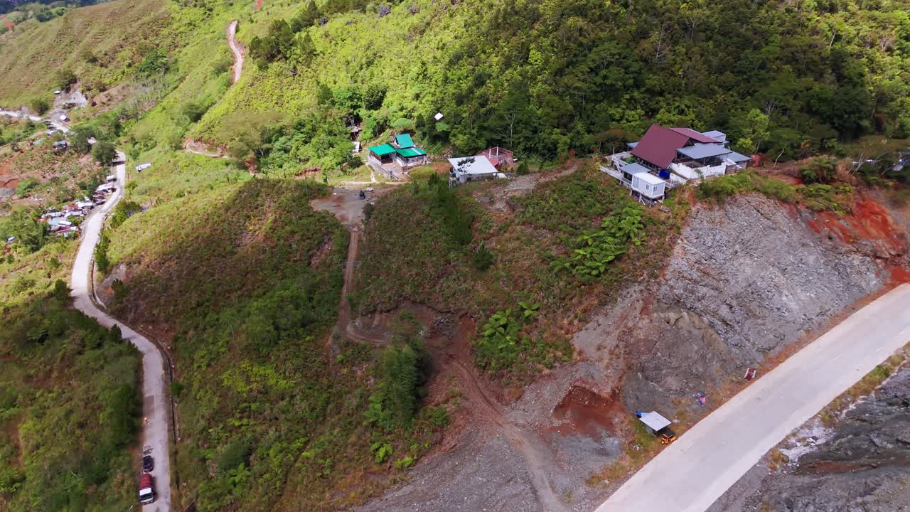 Aerial view of a remote mountain hillside settlement featuring scattered houses, winding rural roads, forest vegetation, and rugged terrain. Scenic landscape showing elevated living areas and natural