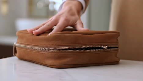 close-up of a hand resting on a brown suede toiletry bag