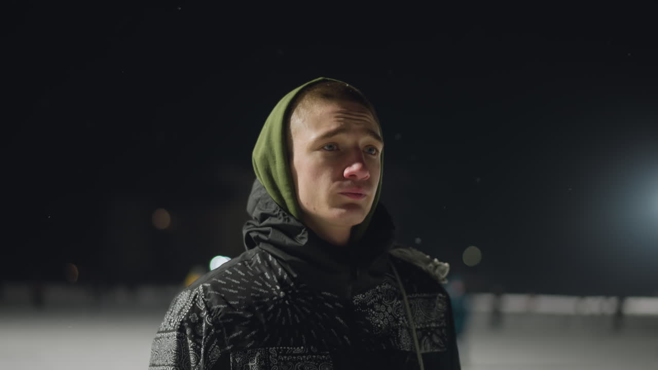unhappy young man walking across snow covered ice rink at night wearing hooded jacket with visible cold breath distant skaters under bright floodlights moody winter scene introspective