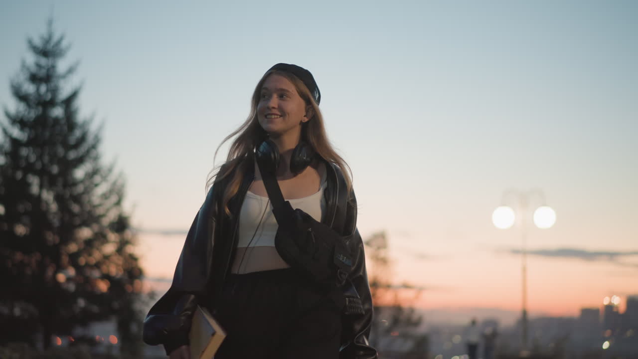 Giovane donna bianca che cammina per strada al tramonto durante l'ora d'oro, con skyline sullo sfondo, berretto, cuffie, libro in mano, sorriso, giacca comoda, luci soffuse del crepuscolo, ritratto spontaneo di una studentessa universitaria che si gode una serata in città