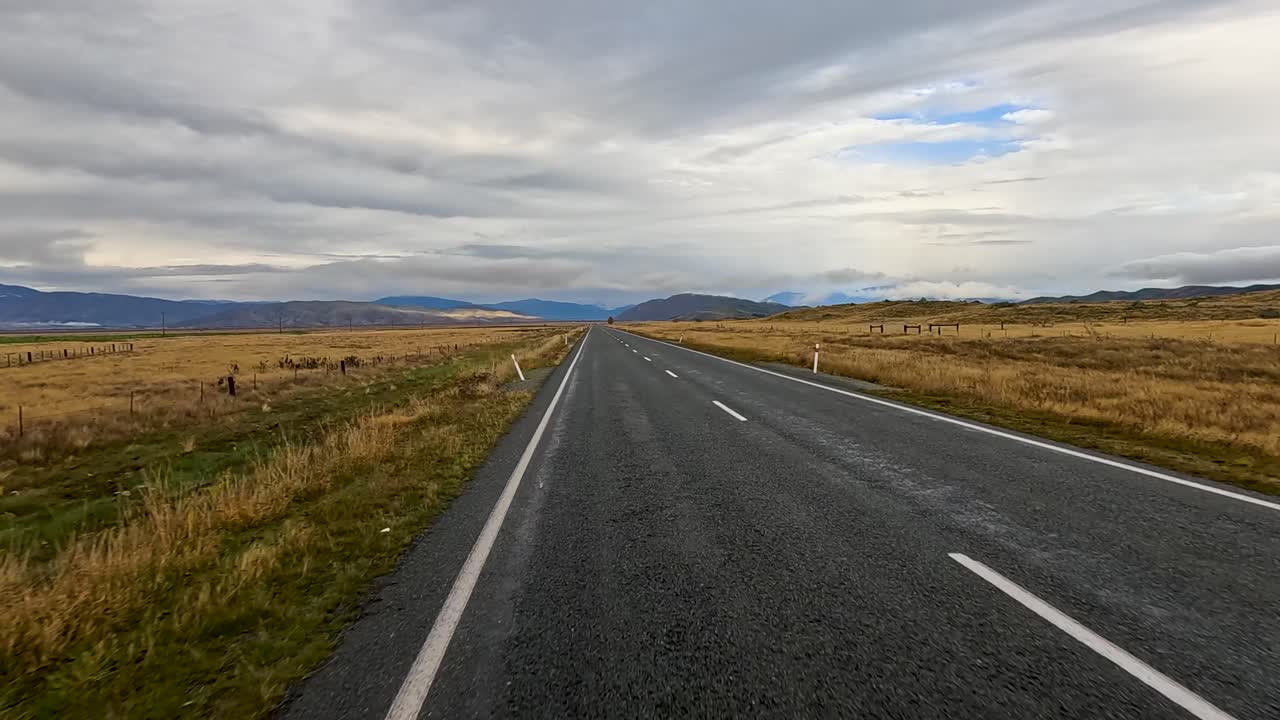 A vehicle travels along a straight, empty rural road through open farmland near Queenstown, New Zealand, under overcast skies with soft natural lighting and steady forward camera movement