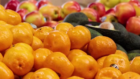 a person picks oranges from a vibrant market display with avocados and apples.