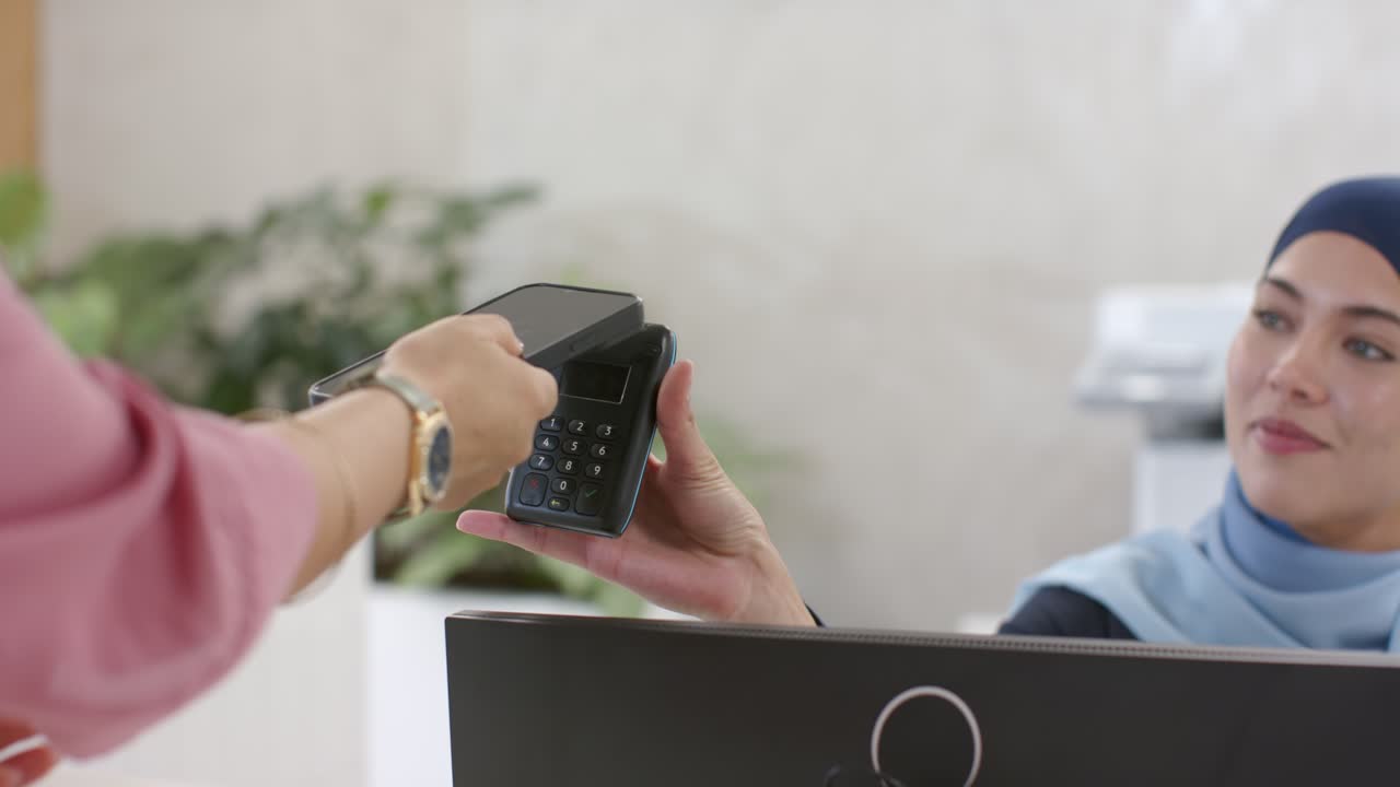 African American receptionist in scarf lifting reader at desk while woman coming to pay with phone