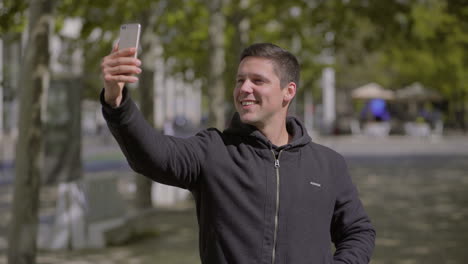 Un-Joven-Sonriente-Tomando-Selfie-Con-Un-Smartphone-Al-Aire-Libre