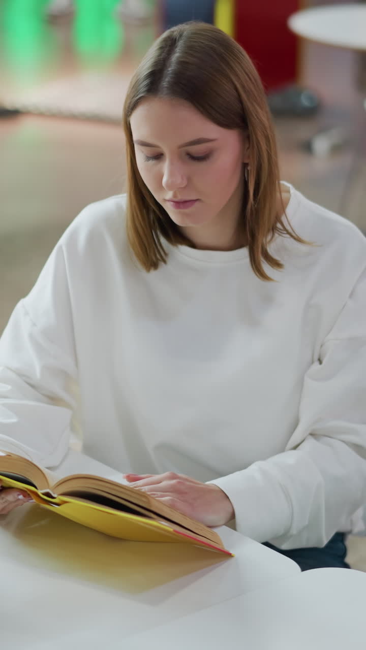 joven leyendo su libro mientras beba café en una mesa en el centro comercial, entorno casual con bolsas de compras cerca, personas caminando en el fondo