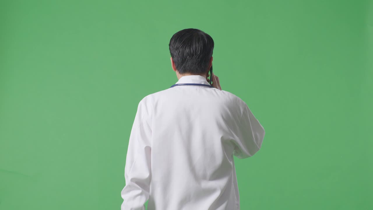 병원 에서 녹색 스크린 배경 에 걸어 다니는 동안 스테토스코프 를 들고 스마트폰 으로 대화 하는 아시아 남자 의사 의 뒷면