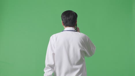 back view of asian male doctor with stethoscope talking on smartphone while walking on green screen background in the hospital