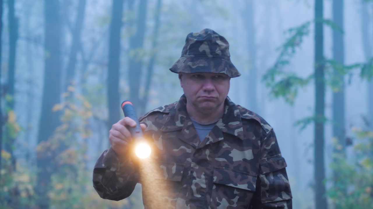 En mand i camouflage går gennem skoven med en lommelygte i hånden i vådt, regnfuldt vejr.