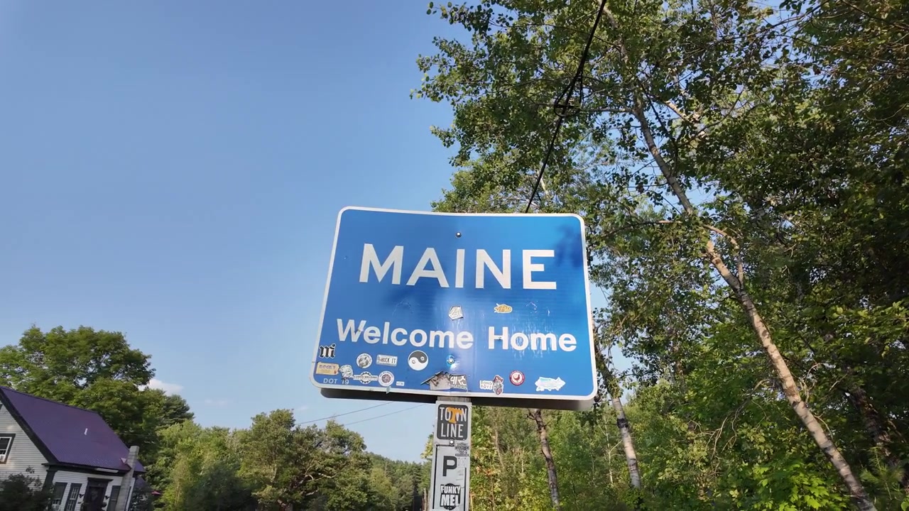 Cedule „Vítejte doma v Maine“ u zalesněné silnice, zatímco kamera panorámuje doleva.