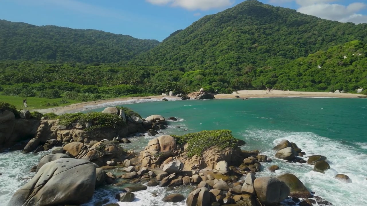 Orbita aerea attorno a massi di granito massicci e una cala turchese a Playa Cañaveral, con alle spalle montagne della giungla nel Parco Nazionale Tayrona, Colombia.