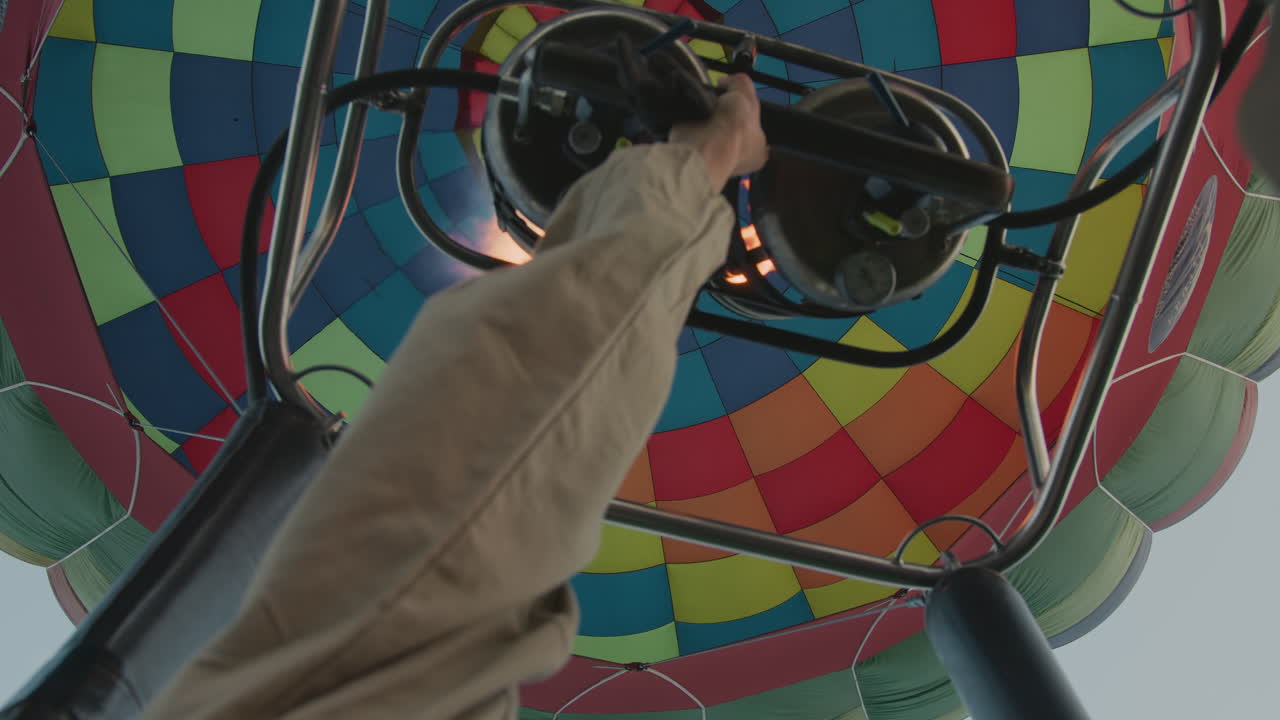 Close up of woman pilot adjusting metal burner handle to increase flame under multicolor balloon canopy against clear sky during sunrise flight creating dynamic aerial adventure scene