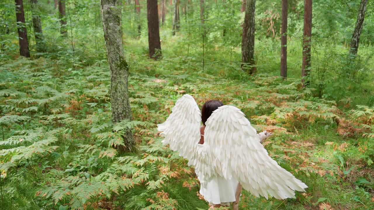 Young woman dressed like ancient goddess Psyche with large white feathered wings walks slowly through lush green forest holding rustic staff, evoking mythological energy