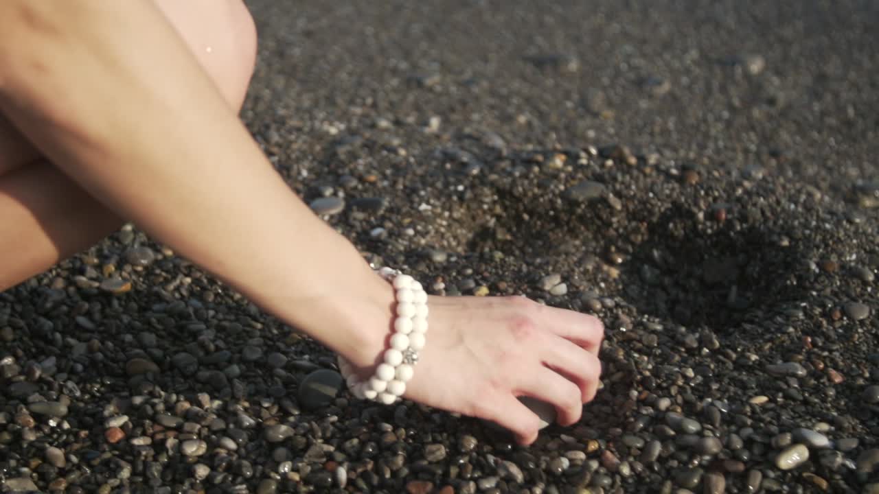 A girl's hand reaches out and picks up a smooth rock on a beach