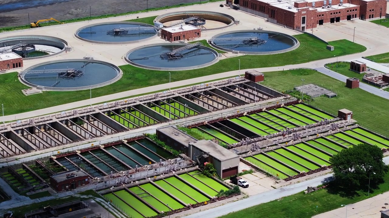 Forward drone shot above a water reclamation facility revealing bright green trickling filter beds, circular clarifiers, and brick operations buildings.