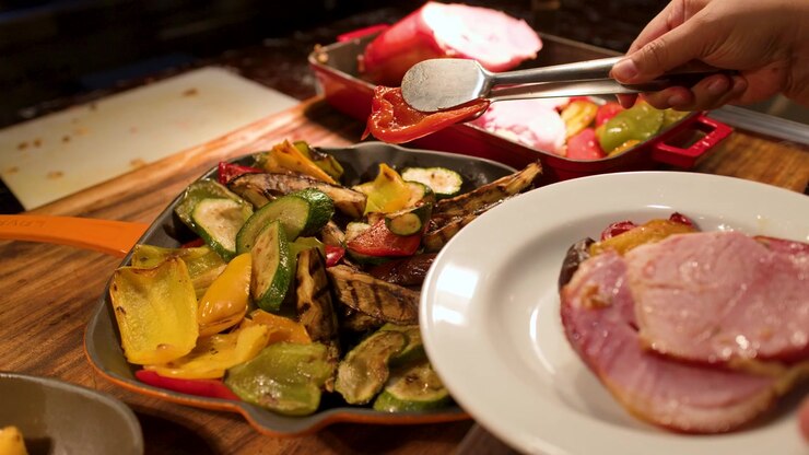 Una persona usa pinzas metálicas para servir pimientos asados en un plato con jamón