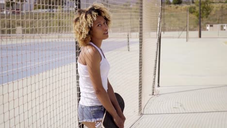 Young-woman-with-skateboard-leans-against-fence