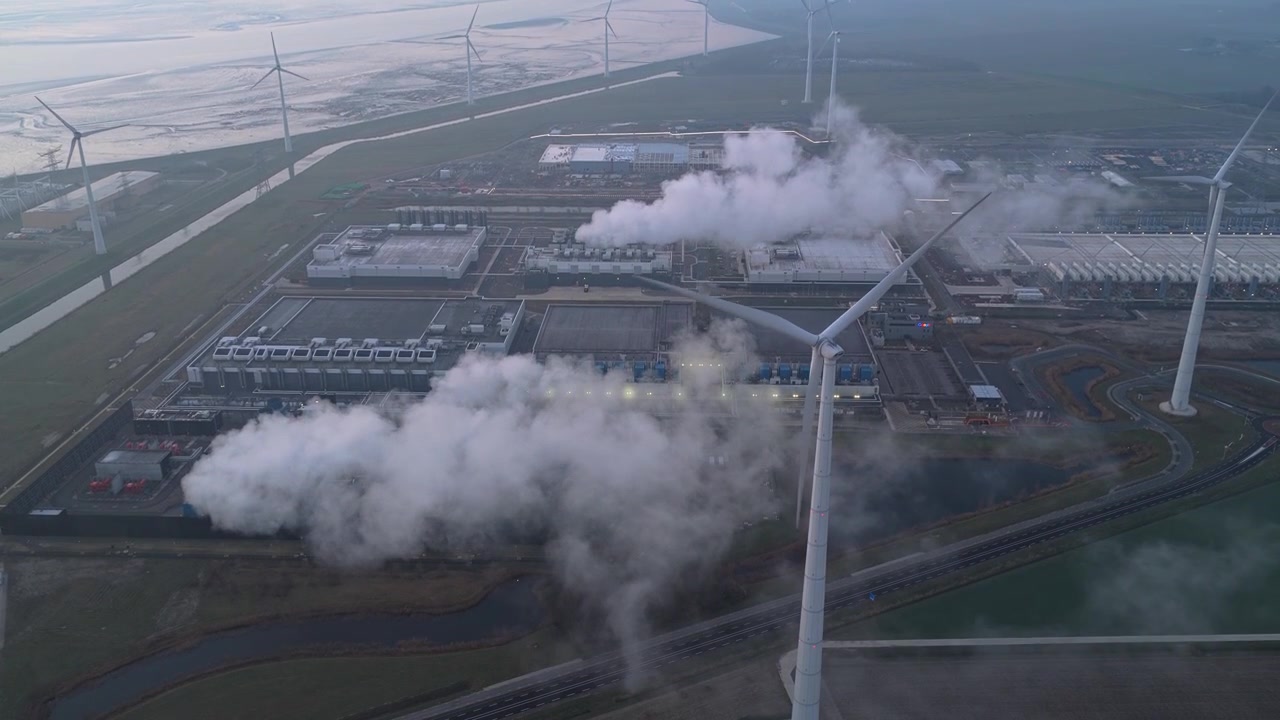 Prospettiva drone del vasto complesso di server farm di Google che rilascia vapore dai sistemi di raffreddamento, circondato da turbine eoliche che generano energia rinnovabile