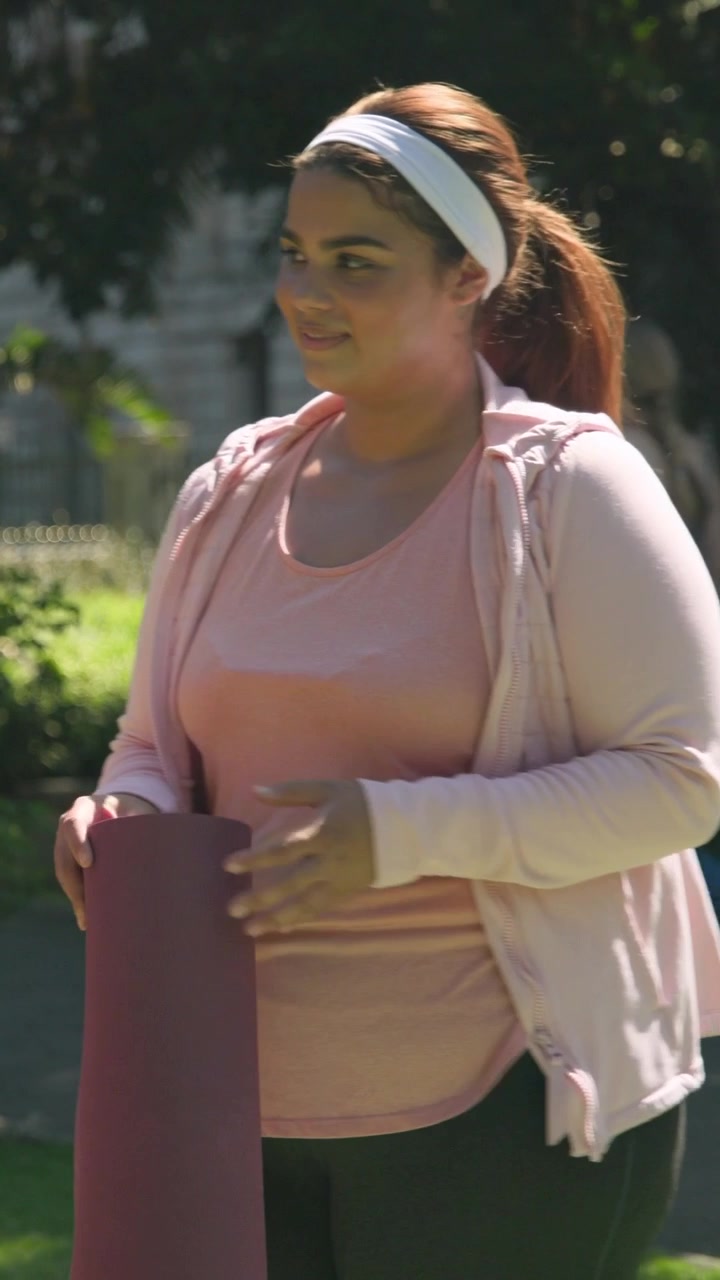 Video verticale: Donna con felpa rosa che entra da destra, portando un tappetino viola su un sentiero del parco, mentre va a lezione.