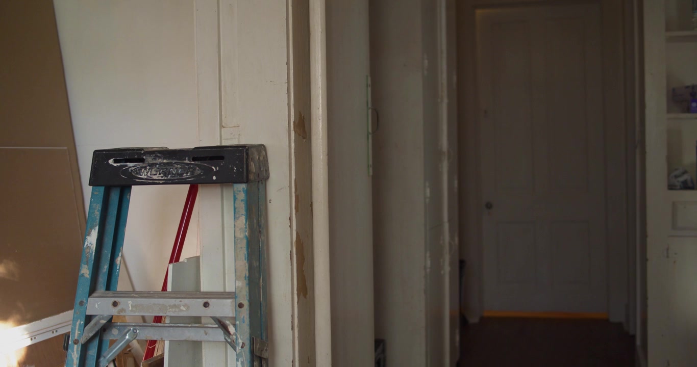 Interior view of a home remodeling project in progress, featuring a blue stepladder near unfinished walls in a hallway leading to a closed door