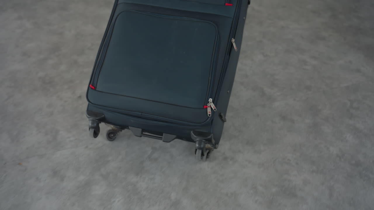 Rear view of man dragging large wheeled travel bag behind him as he enters hotel room with carpeted floor, soft daylight through window, neutral walls and minimal furnishings c